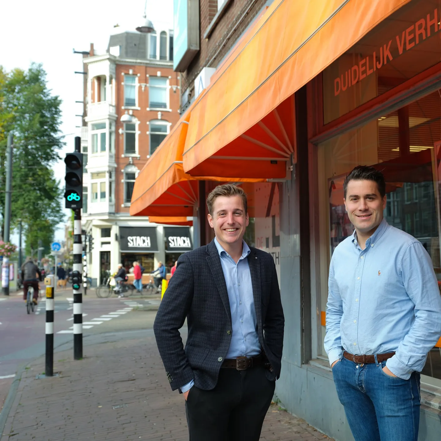 Twee hypotheekadviseurs staan voor de ingang van De Hypotheekshop Overtoom in Amsterdam, onder opvallende oranje zonneschermen.