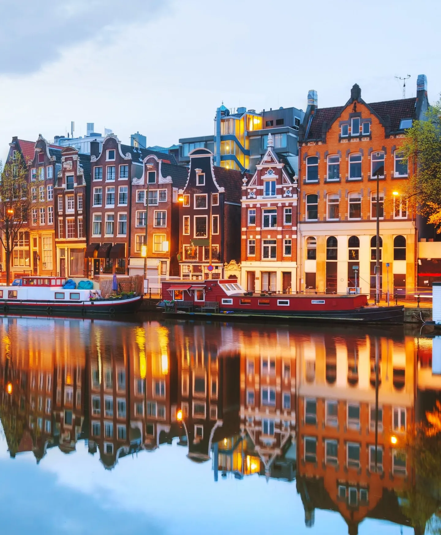 Verlichte Amsterdamse grachtenpanden en woonboten weerspiegelen in het water bij schemering.
