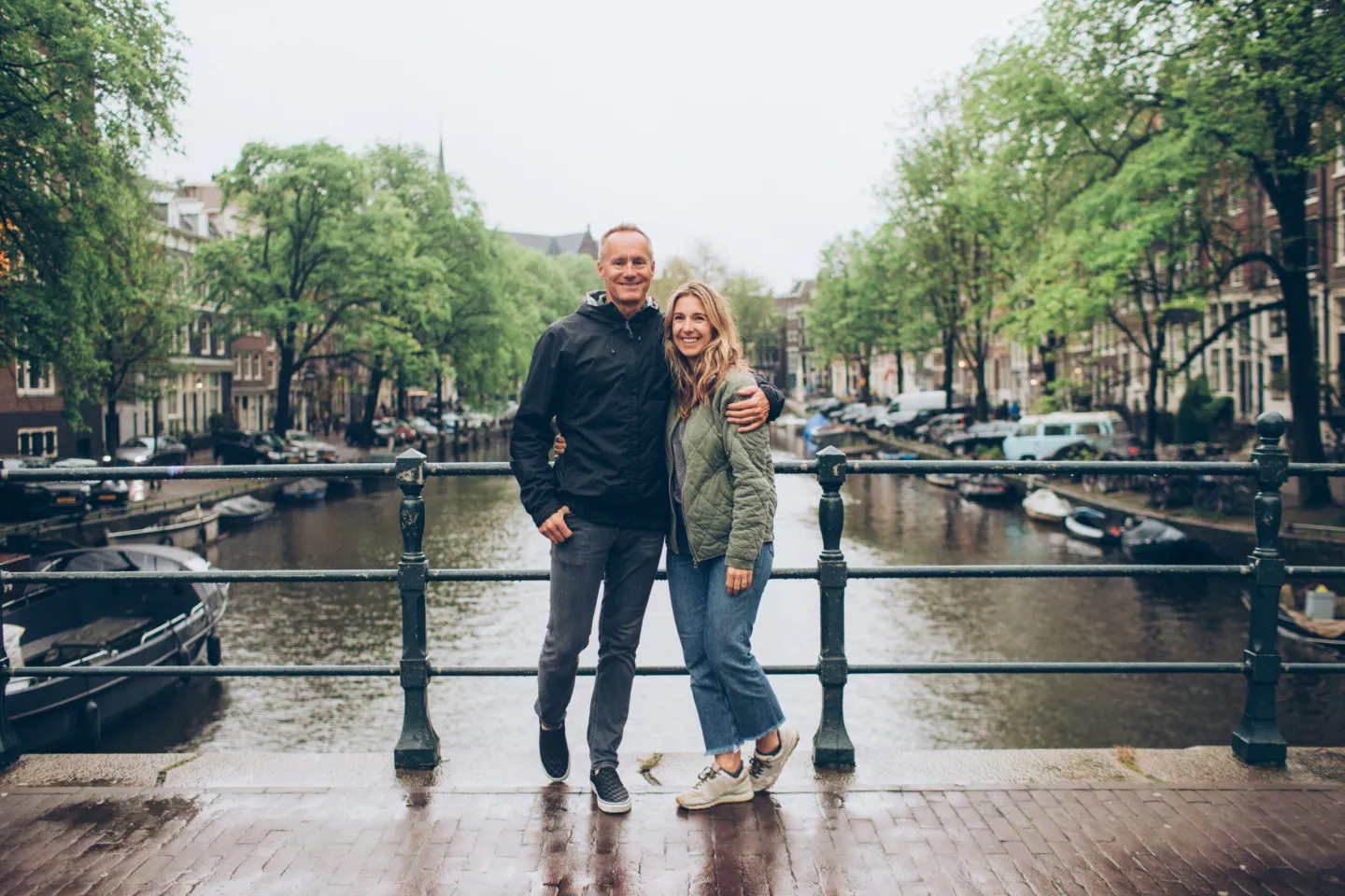 Een stel staat op een brug boven een Amsterdamse gracht, met woonboten, bomen en grachtenpanden op de achtergrond.