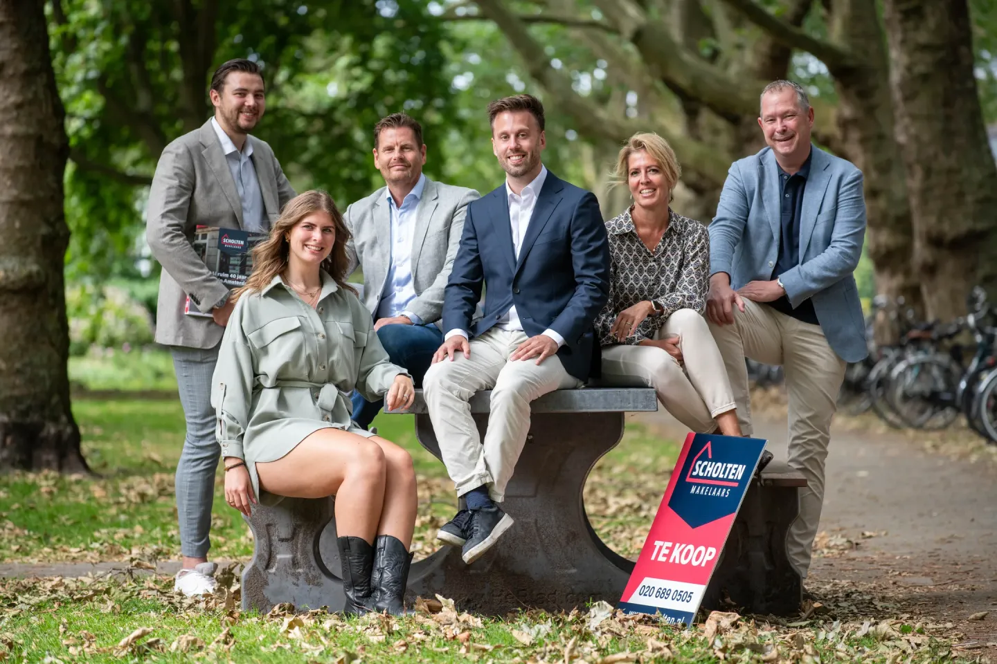 Groep medewerkers van Scholten Makelaardij poseren bij een bankje in een groen park, naast een bord met de tekst ‘Te koop’.