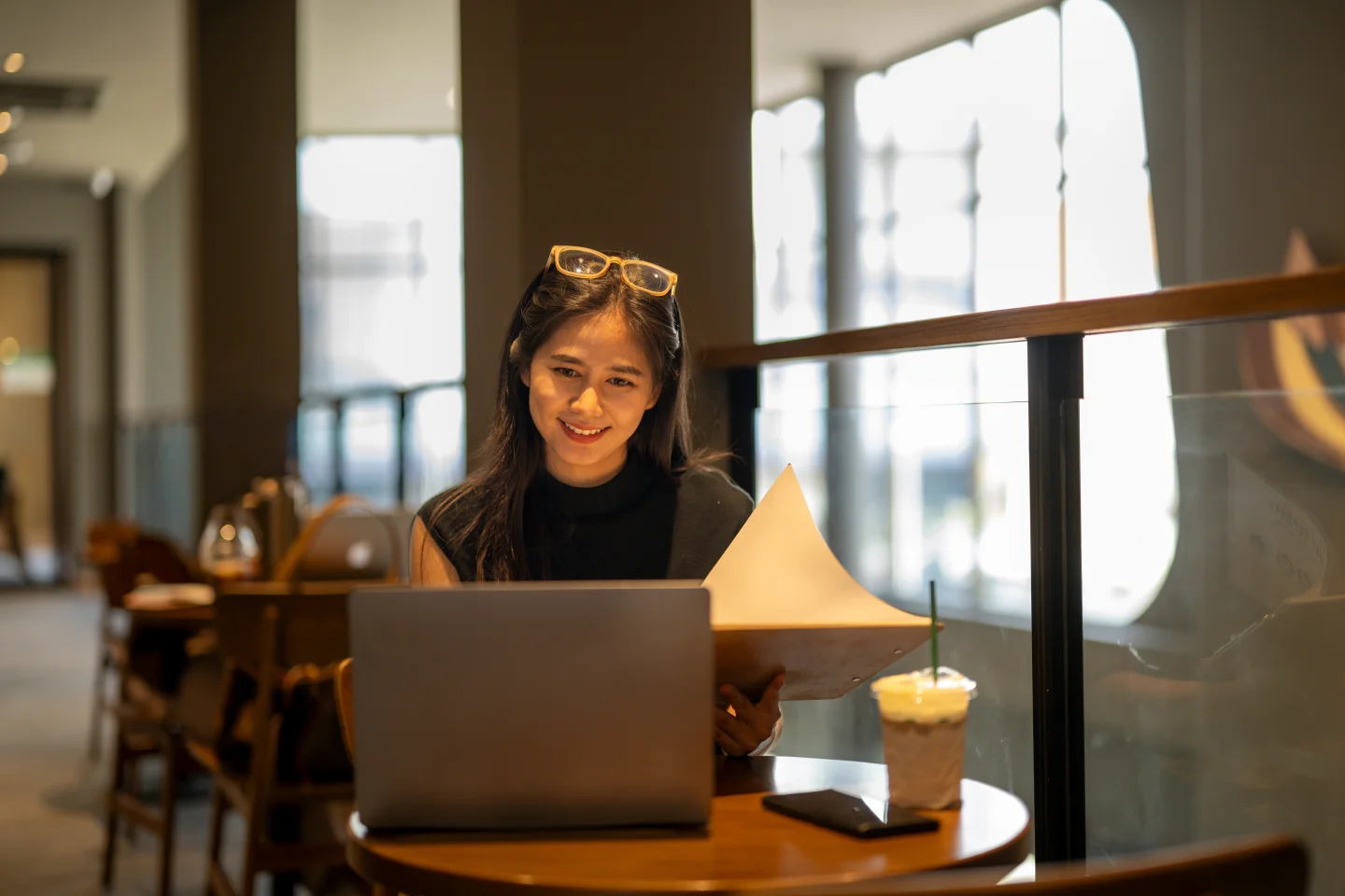 Jonge vrouw glimlachend aan het werk in een café, gebruikt haar laptop en heeft documenten vast.