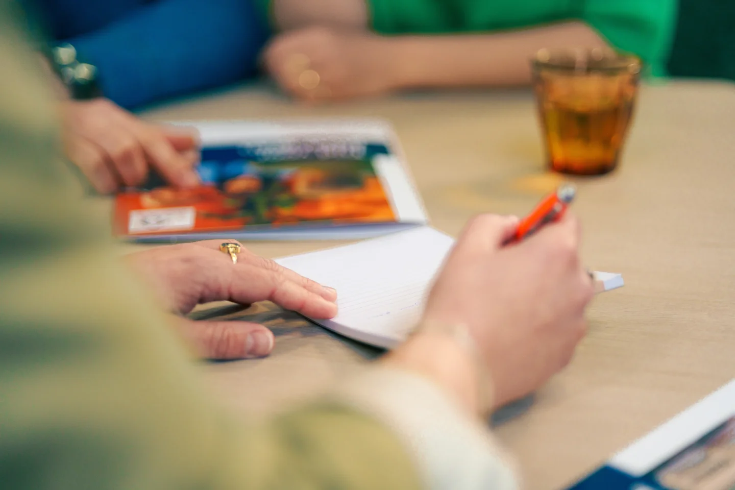Persoon schrijft met een pen in een notitieblok tijdens een overleg aan tafel, met anderen op de achtergrond.
