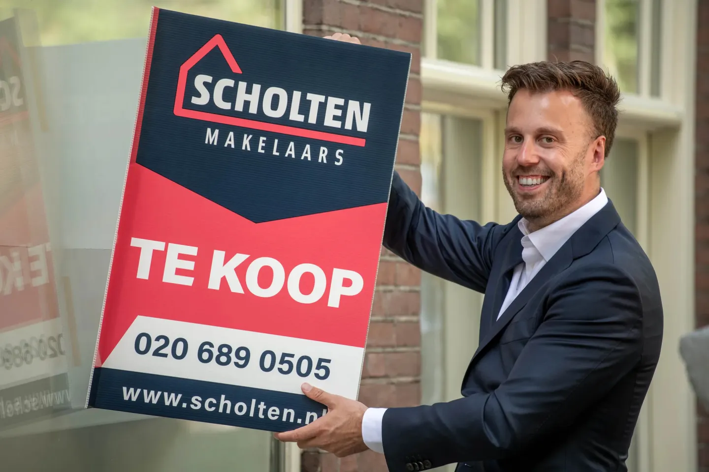 Man in pak houdt een Scholten Makelaars-tekoopbord vast voor een woning in Amsterdam.