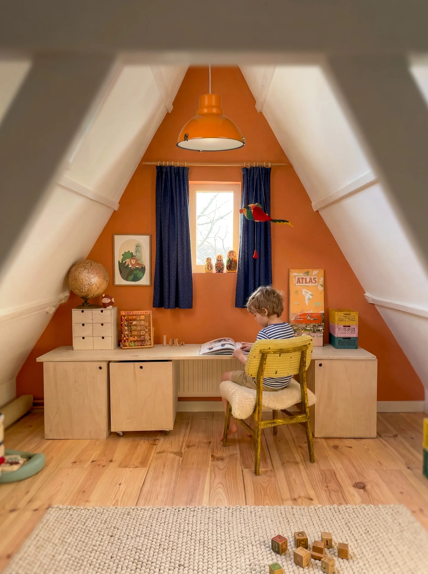 Kind leest aan een bureau in een lichte kinderkamer met oranje wand, houten vloer en speelgoed op de grond.